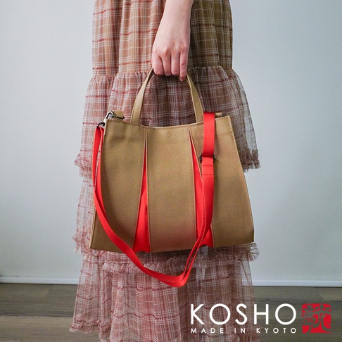 米色棉帆布扇形托特包，搭配鮮豔的紅色織帶提把與可拆卸斜背帶，包身有紅色飾條點綴。