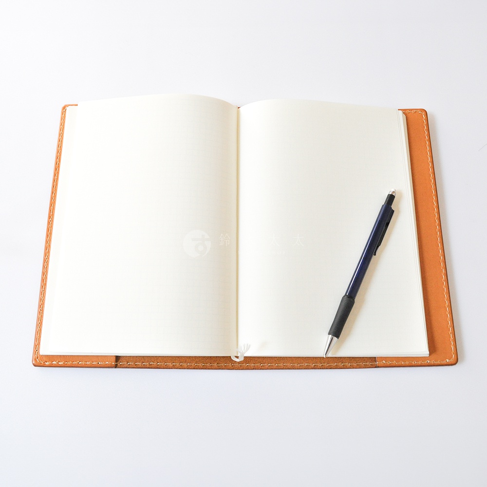 一個攤開的日本HERZ職人手作皮革筆記本書套，採用溫潤的淺棕色Latigo軟皮革製成，邊緣有細緻的車縫線工藝，中間放置著一本展開的空白筆記本與一支深藍色原子筆。