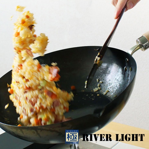 黑色金屬炒鍋，鍋內有炒飯飛濺，由木製鍋鏟翻炒，鍋柄為金屬與木製結合。
