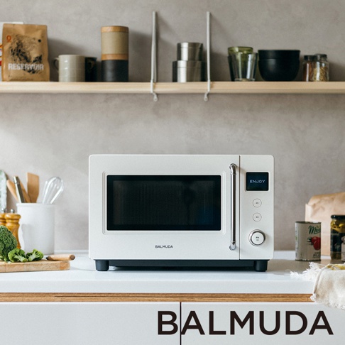 位於廚房白色檯面上的白色BALMUDA微波爐，採正面視角拍攝。機身為簡約方正設計，右側設有金屬拉門把手、電子螢幕與旋鈕控制，螢幕顯示ENJOY字樣。背景為淺灰色牆面與安裝在牆上的木質層架，層架上擺放著馬克杯、金屬罐與玻璃杯等廚房用品。左側檯面有裝著料理工具的白色容器及木製砧板與綠色花椰菜，右側檯面放有罐裝食品。圖片下方標註有BALMUDA字樣。