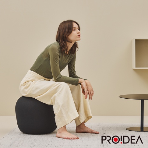 一位女性坐在一顆黑色的針織彈力平衡球上，身穿綠色長袖上衣和米白色寬褲，背景為淺米色牆壁和簡約的室內陳設。