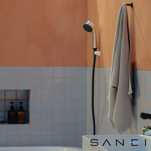 銀色金屬蓮蓬頭，黑色軟管，掛在橘色牆壁上的灰色紋理毛巾，下方為淺藍色磁磚牆面，壁龛內有兩個深琥珀色按壓瓶。