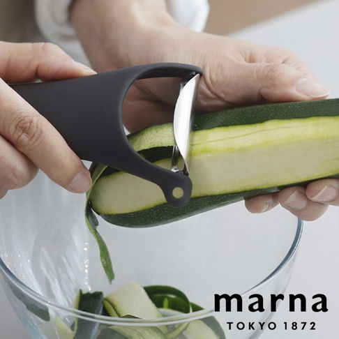 一名使用者手持黑色的 marna 多功能削皮器，正在將一顆綠色櫛瓜削成薄片，削下的皮片落入下方的透明玻璃碗中，畫面呈現簡約的廚房用品使用情境。