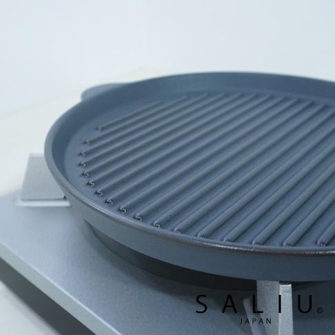 The chef 多功能耐熱陶製萬用波浪圓烤盤 S，呈現深灰色，具有規則的波浪烤紋，圓形盤面，兩側有提把，整體為陶製材質，置於淺灰色爐架上。