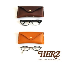 兩個皮革眼鏡收納盒，上方為棕色，下方為橘色，分別搭配一副黑色方框眼鏡和一副圓框眼鏡。右下角有HERZ品牌標誌。