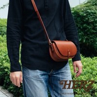 職人手作皮革迷你馬鞍包，採用棕色皮革製成，圓弧造型，附有細長肩帶，單肩斜背於身穿黑色襯衫和牛仔褲的模特兒身上。