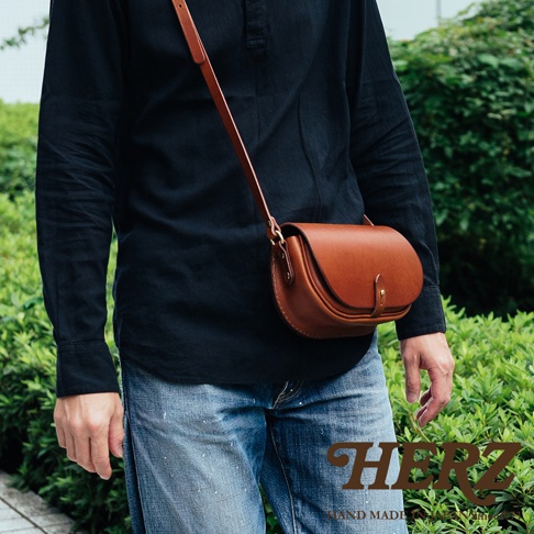 職人手作皮革迷你馬鞍包，採用棕色皮革製成，圓弧造型，附有細長肩帶，單肩斜背於身穿黑色襯衫和牛仔褲的模特兒身上。