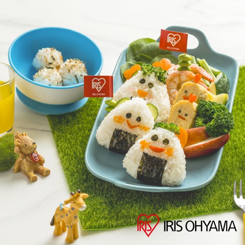 藍色餐盤上擺放著可愛的動物造型飯糰，搭配蝦子、花椰菜、紅蘿蔔、玉子燒和蘋果切片，飯糰上插有印有「IRIS OHYAMA」字樣的紅色小旗子，旁邊還有裝有飯糰的藍白色碗以及裝有橘色飲品的玻璃杯。