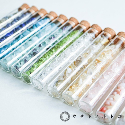 一系列玻璃試管瓶，內含不同顏色和大小的礦石顆粒，從深藍、淺藍、綠色、透明、黃色到粉紅色，瓶口均以軟木塞封閉。