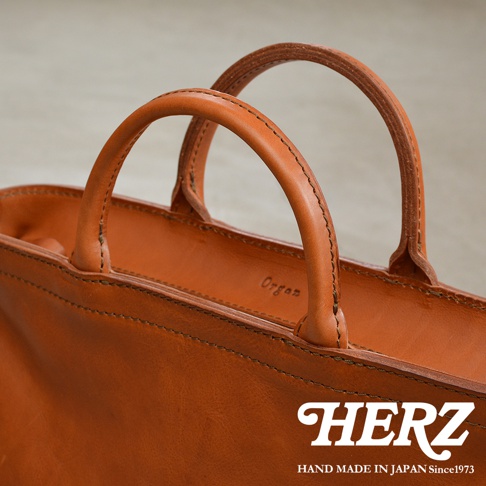 HERZ 品牌手工皮革托特包，特寫其焦糖色皮革手柄及縫線細節，「Organ」品牌字樣隱約可見。