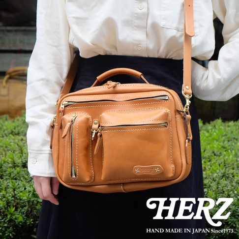 一款棕褐色皮革製作的兩用單肩包，具有多個拉鍊口袋和一個翻蓋口袋，包身上有品牌標誌，由職人手工製作。