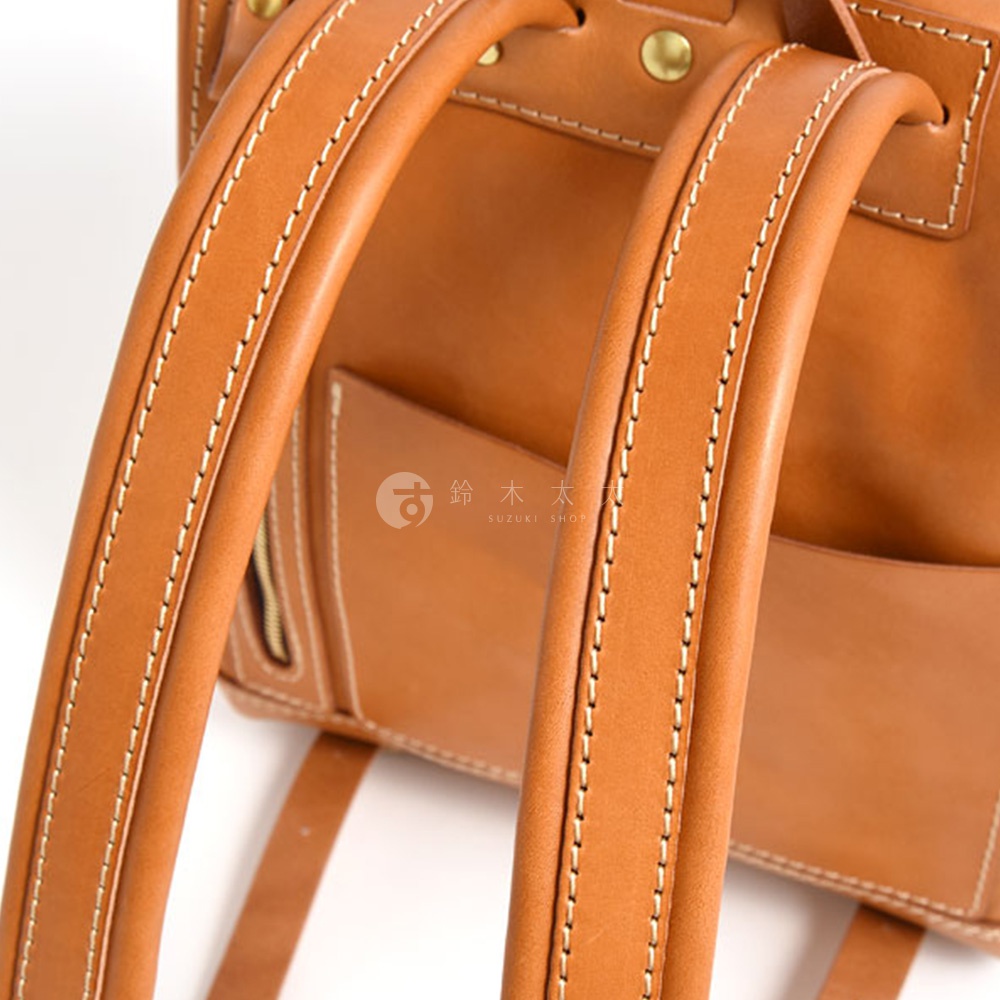 職人手作皮革雙口袋折蓋後背包，特寫縫線精緻的棕褐色皮革肩帶和背包主體，可見金屬鉚釘和拉鍊細節。