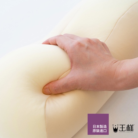 手壓在淺黃色、柔軟的長條形抬腿枕上，枕頭呈現出凹陷的形狀。枕頭表面為光滑布料，縫線清晰可見。角落有紫底白字的「日本製造 原裝進口」及「王樣」品牌標誌。