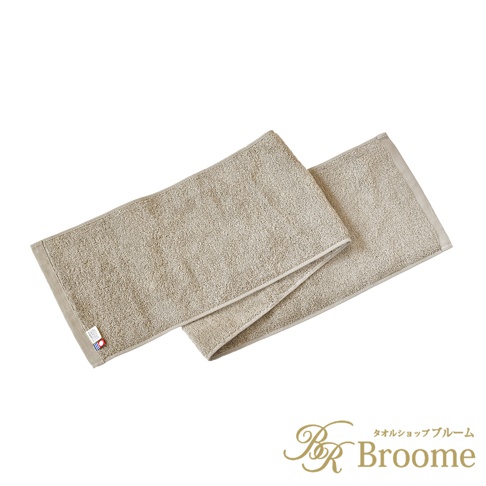 兩條淺米色長毛巾，材質為長毛絨，質地柔軟。其中一條毛巾疊在另一條上方，邊緣有細緻的縫線，左側角落處有一個帶有日文標誌的紅色小標籤。