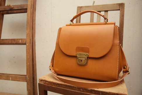 職人手作的淺棕色皮革兩用單肩包，帶有黃銅色鎖扣和縫線細節，整體風格簡約。