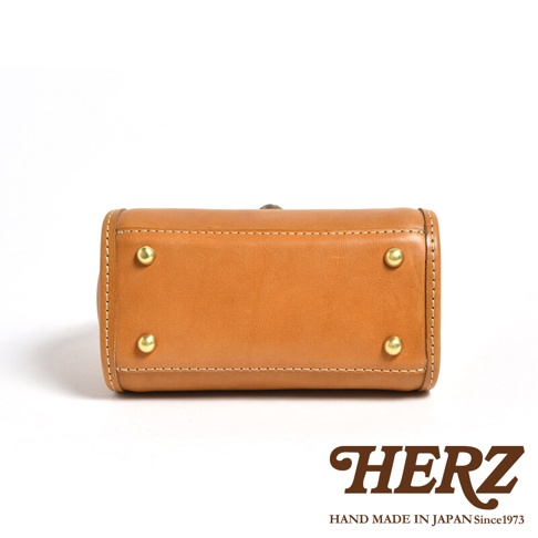 職人手作皮革迷你手提包，呈現溫潤的焦糖棕色，包身結構紮實，頂部飾有金屬旋釦，四個角落設有金色銅釘，整體以白色縫線勾勒出簡潔的輪廓，展現皮革的自然質感。