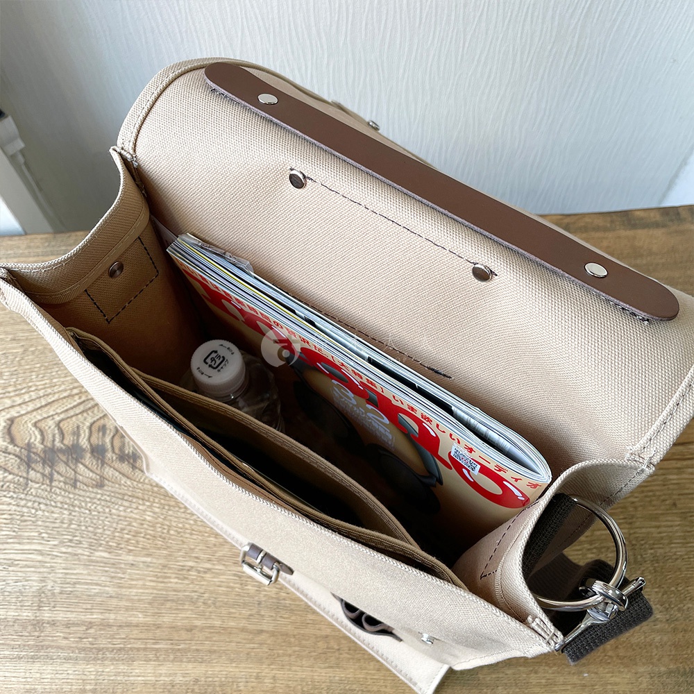 淺米色純棉帆布製成的直式側背包，打開蓋子，內部裝有雜誌、水瓶和文件夾。包包帶有金屬扣和鑰匙圈。