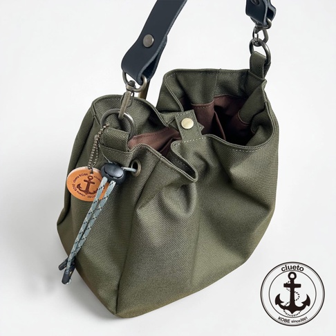 這是一款神戶clueto品牌的BALON mini多用途手提包，選用橄欖綠色的防潑水帆布製成，搭配黑色皮革背帶與金屬掛扣。包身設有束口設計，並配有一個印有錨圖案與clueto KOBE字樣的圓形皮革吊飾，整體呈現簡約實用的日系風格。