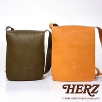日本 HERZ 品牌，展示兩個職人手作皮革軟皮直式斜背包，一個為橄欖綠色，另一個為橘黃色，包包正面皆有品牌字母 Z 的壓印。