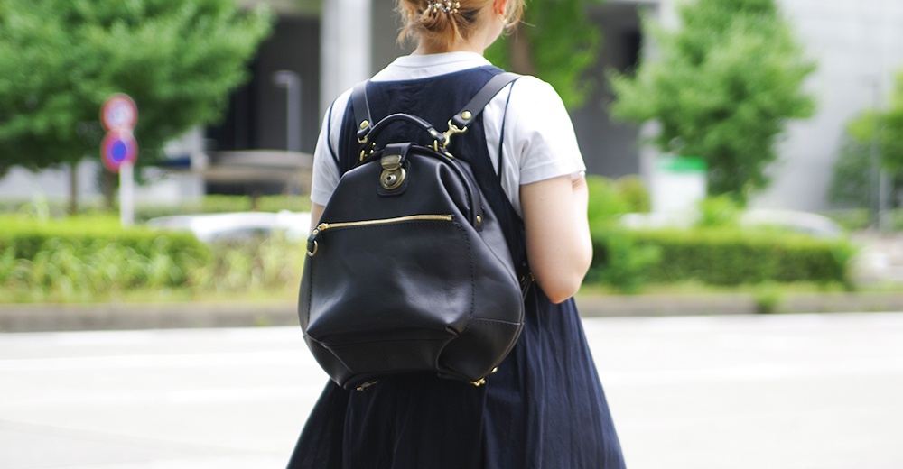 黑色皮革後背包，設有金色拉鍊和五金配件，包身線條簡潔，底部有四個金屬腳釘。