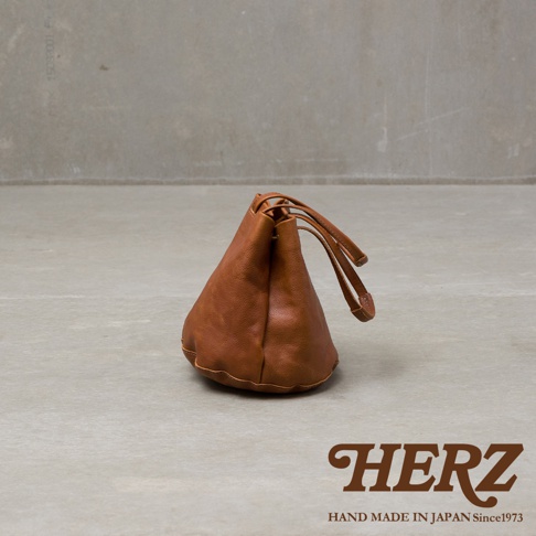 一個日本品牌 HERZ 的棕色手工皮革抽繩包，採用簡約的托特包設計，呈現出獨特的圓錐狀外觀與細膩皮革質感，包袋邊緣有精緻縫線細節，底部呈現立體圓弧形，底部右下方印有 HERZ 品牌名稱與手作產地說明。