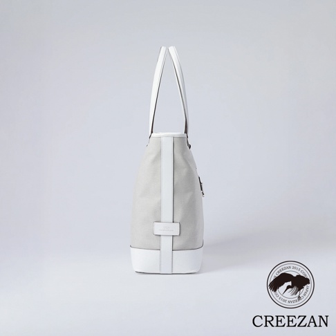 側拍視角的 CREEZAN 品牌大托特包，包身選用灰色質感帆布面料，搭配白色皮革底座、邊條與手提把，整體設計簡約優雅，右下角印有品牌名稱與展翅鶴鳥標誌。