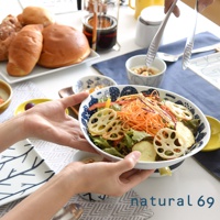 一雙手捧著一個白色陶瓷沙拉碗，碗內裝有豐富的沙拉，包含生菜、紅蘿蔔絲、甜椒、地瓜片和蓮藕片，碗邊有藍色花紋。背景有多種麵包、小碟和餐具。