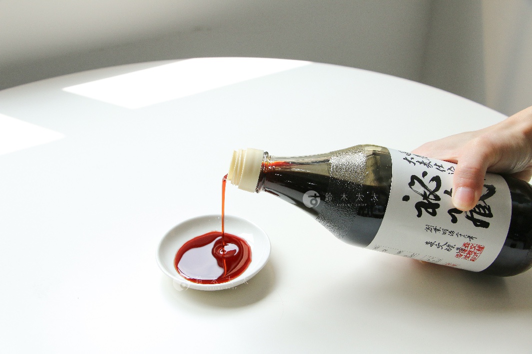 手持玻璃瓶裝醬油，將深褐色醬油傾倒入白色小碟中，醬油呈現濃稠狀。