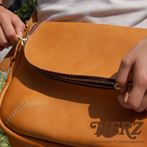 HERZ 職人手作皮革大容量馬鞍包，展示了精緻的縫線細節和高品質的皮革質感，拉鍊拉合處的皮革拉片和金屬拉環清晰可見，標有「HAND MADE IN JAPAN Since1973」的品牌標誌。