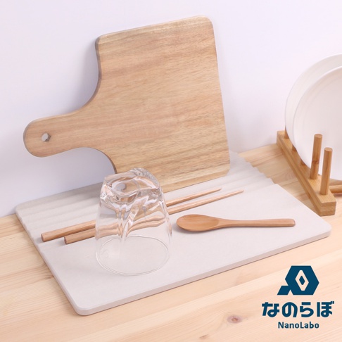 淺灰色珪藻土置物墊上擺放著一個透明玻璃杯、兩支木筷和一支木湯匙，後方則有一個木質砧板和一個竹製碗盤架。