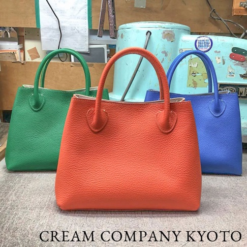 三個顏色鮮豔的皮革手提托特包，從左至右分別為綠色、橘紅色和藍色，包身帶有紋理，提把設計圓潤。