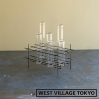 黑色金屬支架，帶有六個透明玻璃試管狀花器，置於深色桌面，背景為淡黃色牆面。