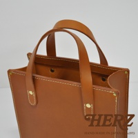 職人手作皮革直式托特包，採用天然皮革材質，呈現溫潤的焦糖棕色。包款設計簡潔，有兩個皮革提把，以白色縫線和金色銅釦裝飾，整體風格樸實耐用。