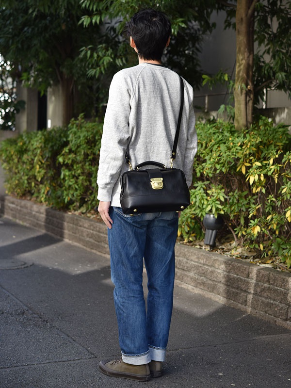 黑色皮革迷你口金單肩包，搭配金色金屬扣環和可拆卸肩帶，由一名穿著灰色運動衫和牛仔褲的男子側身展示。