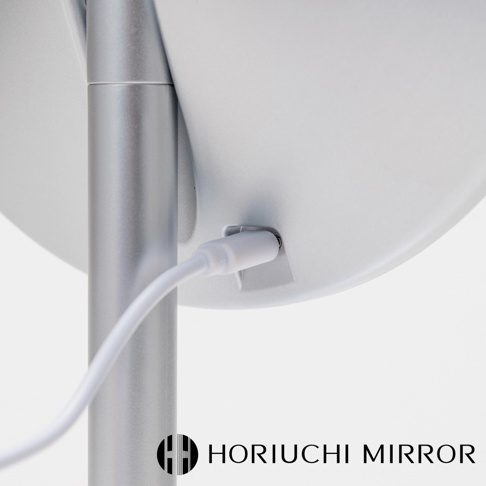 銀色金屬支架連接至白色圓形化妝鏡背面，一條白色充電線插入鏡背右側的充電孔。