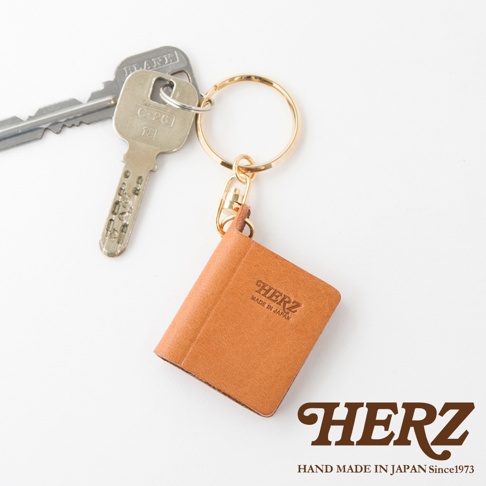職人手作皮革書本造型鑰匙圈，焦糖色皮革，壓有「HERZ MADE IN JAPAN」字樣，搭配金色鑰匙圈與銀色鑰匙。