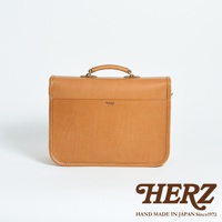 焦糖色皮革手提包，方形設計，頂部有提把和金屬扣環。包身正面有一個壓印的品牌標誌，縫線精緻，質感厚實。