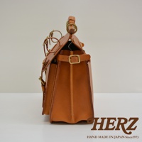 側面展示一款棕褐色皮革旅行包，包身結構方正，配有黃銅色金屬扣環和皮帶。包側裝飾有雙層皮質邊條，手工縫線清晰可見，提把由木質和皮革結合而成，整體呈現復古風格。