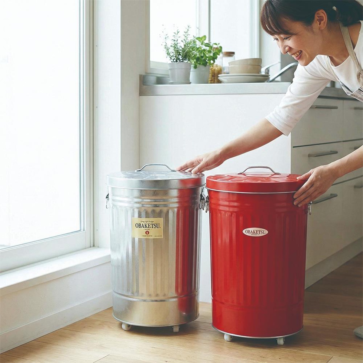 以手工製作的 OBAKETSU 鍍鋅鋼製附輪垃圾桶，其中一個為銀色，另一個為紅色，在明亮的廚房裡。