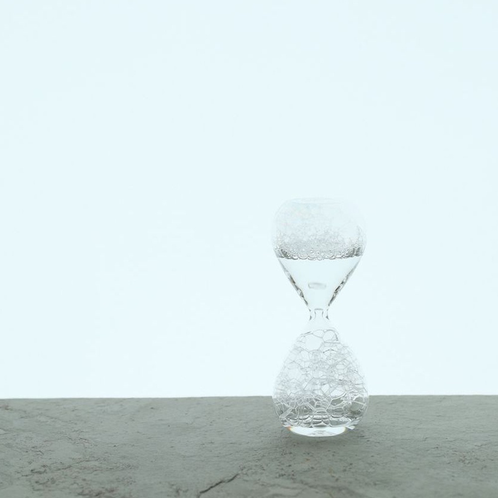 awaglass 泡時計，透明玻璃材質，沙漏形狀，上半部裝有水，水面有許多細小的氣泡，下半部也充滿了氣泡，置於灰色石質表面上，背景為淺藍色。