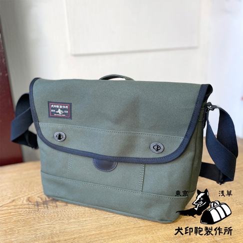 這款黑犬印純棉帆布雙扣郵差包，採用軍綠色帆布材質，配有深色滾邊與雙扣設計。包包有可調整的肩帶，正面有一個紅色標籤，上面印有日文文字與狗狗圖案。