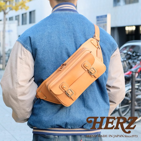 棕色皮革製作的斜背包，斜挎於身穿牛仔外套的男士背部，包身有兩個翻蓋口袋，均以雙釦設計固定，展現職人手作質感。