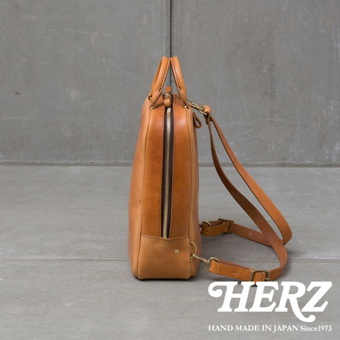 職人手作皮革Organ商務後背包 M-10，採用自然光澤的棕色皮革製作，側邊視角展示其簡約俐落的設計，搭配金屬拉鍊與可調整的皮質肩帶，底部有品牌HERZ標誌和HAND MADE IN JAPAN Since1973字樣。