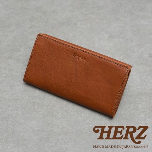 這款日本 HERZ 的 Organ 職人手作皮革長夾呈現焦糖棕色，採用質感細膩的皮革材質製作，正面印有燙印字樣，整體外型簡約洗鍊，以俯視角度展現其精緻的縫線細節與皮革光澤，右下方並標示著 HERZ 的品牌標誌與產地資訊。