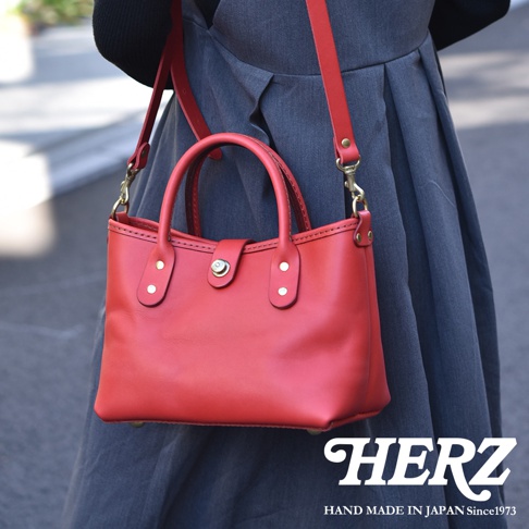 紅色皮革迷你托特包，附有手提把與可拆卸肩帶，包身有金屬扣與鉚釘裝飾，由日本職人手工製作。