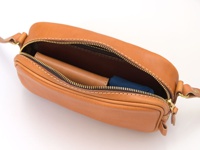 職人手作皮革隨身單肩小包，呈現自然棕色，材質為皮革，包身拉鍊開啟，內部可見一個深棕色皮夾和一個藍色摺疊傘。