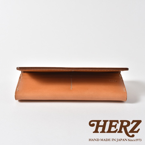 一個呈現皮革原色的日本HERZ品牌職人手作長夾，採用簡約的翻蓋設計與結實的縫線工藝，從略微俯視的角度展示其細緻的皮革紋理與內部結構，右下角印有HERZ品牌標誌與手工製造資訊。