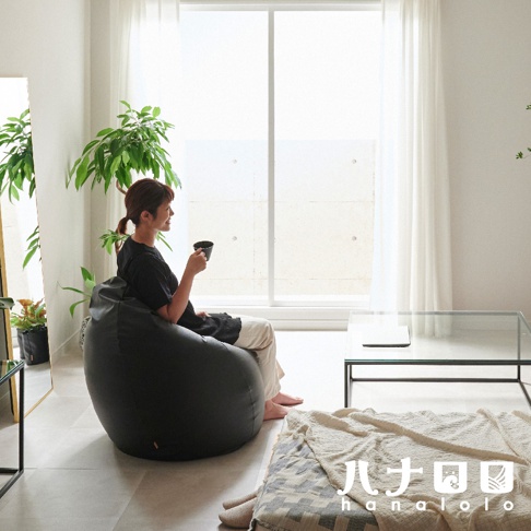 黑色皮革款懶骨頭沙發，坐著一位女性，她手持咖啡杯，身旁有綠色植物和落地窗。