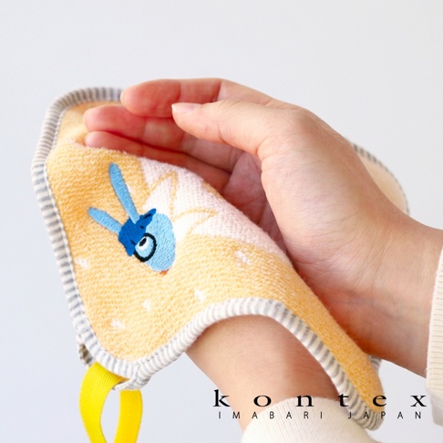 這是一張特寫鏡頭，一隻手輕柔地拿著一塊黃色底色的 Kontex 今治純棉方巾，方巾邊緣帶有灰色條紋飾邊並縫有一個亮黃色的掛環，毛巾表面繡有藍色兔子圖樣，展現出柔軟親膚的質地。