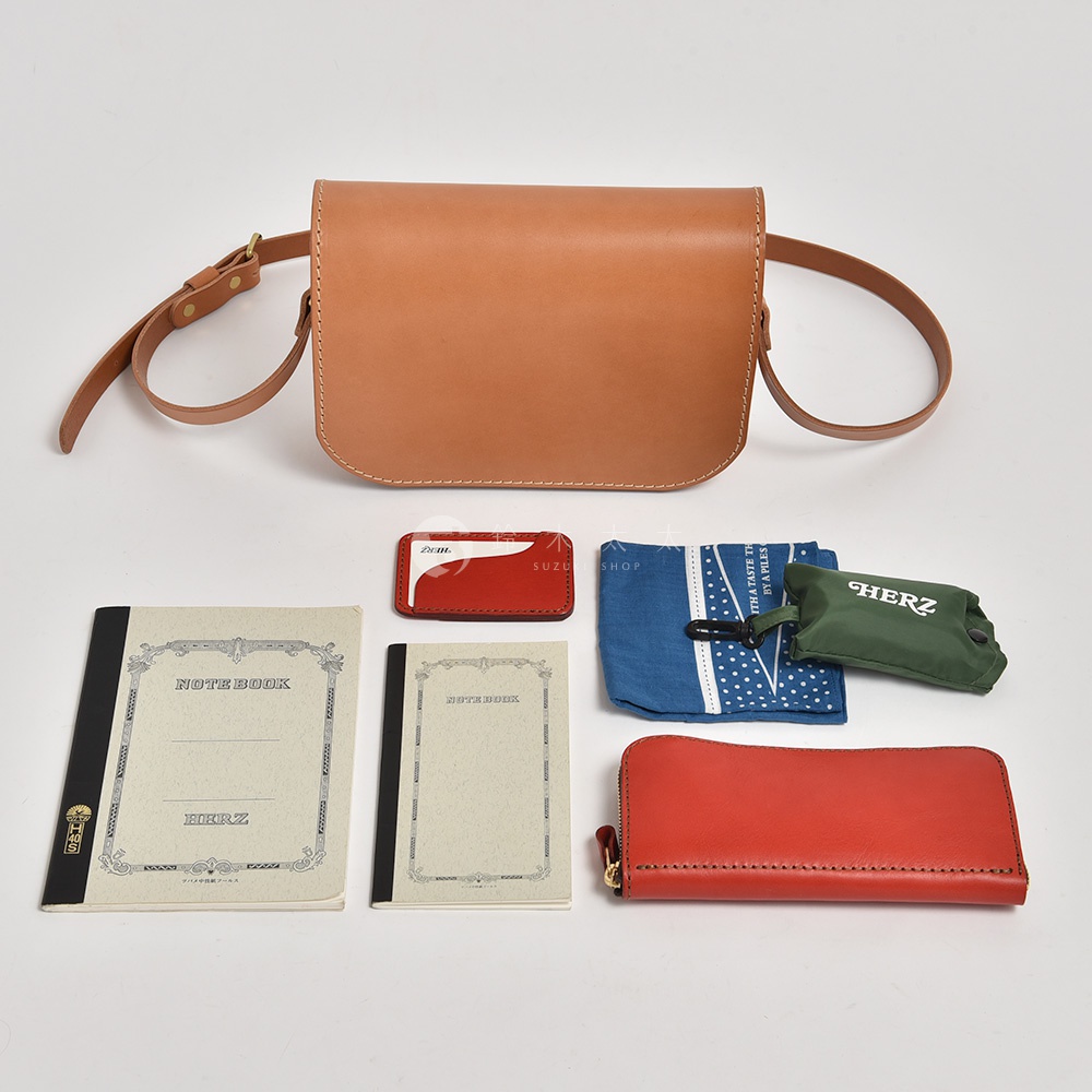 職人手作皮革翻蓋橫式單肩包，淺棕色皮革。配件包含：紅色卡夾、藍色印花布、綠色收納袋、兩本筆記本、紅色長夾。